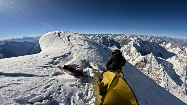 A cliber by a tiny tent placed on a sharp snowy summit. 