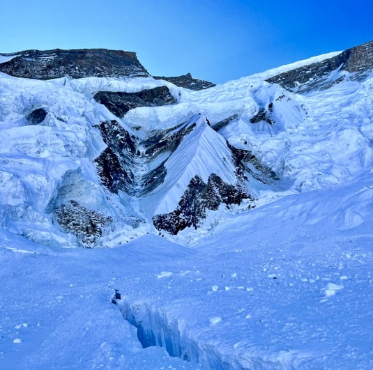 A photo from winter Annapurna I 2024, with a crevasse in the foreground and behind, the Great Couloir, with serac debris falling from the top.