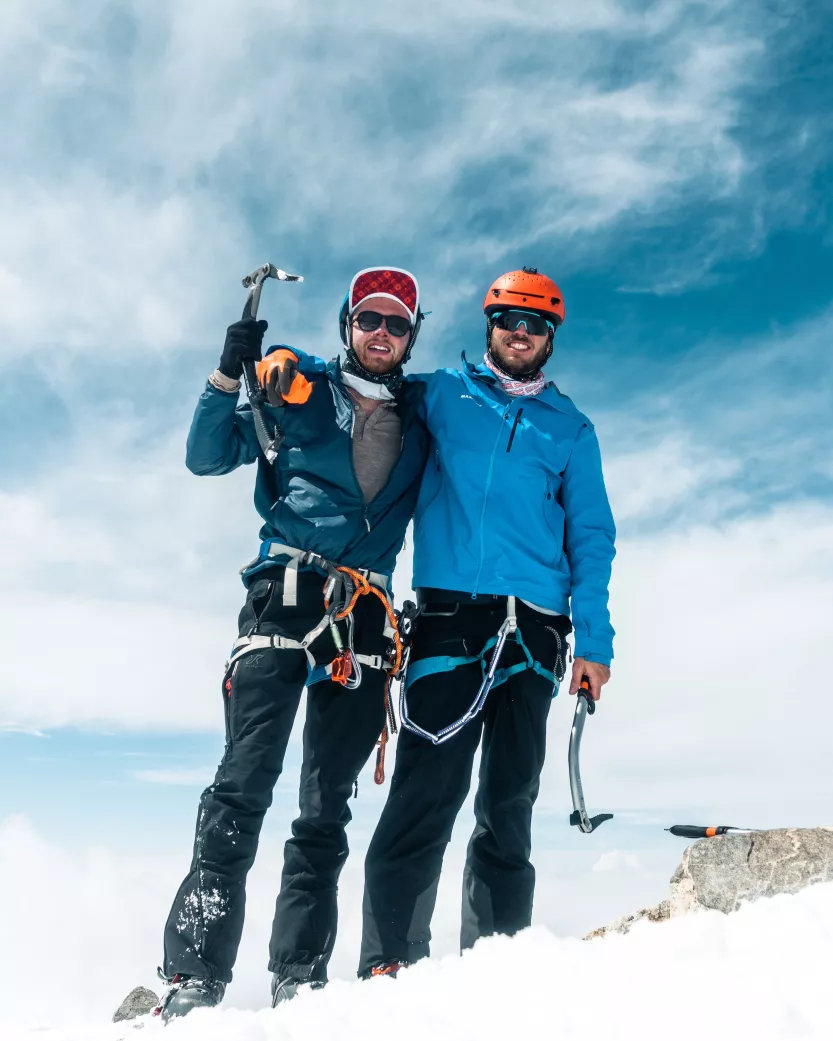 Two men are positioned at the summit of a mountain holding ice axes.