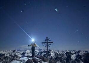 A climber on a summit at night under the starts, with a headlamp