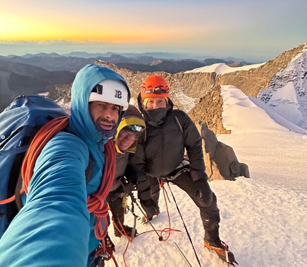 A climber takes a selfie of his group on a snowy summit at sunset