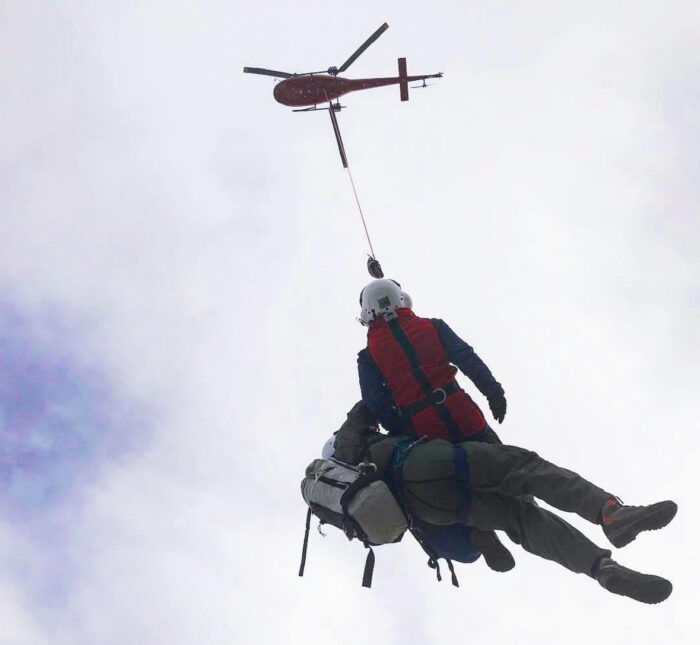 A rescuer hangs from a helicopter by a cable while carrying a patient attached to his back