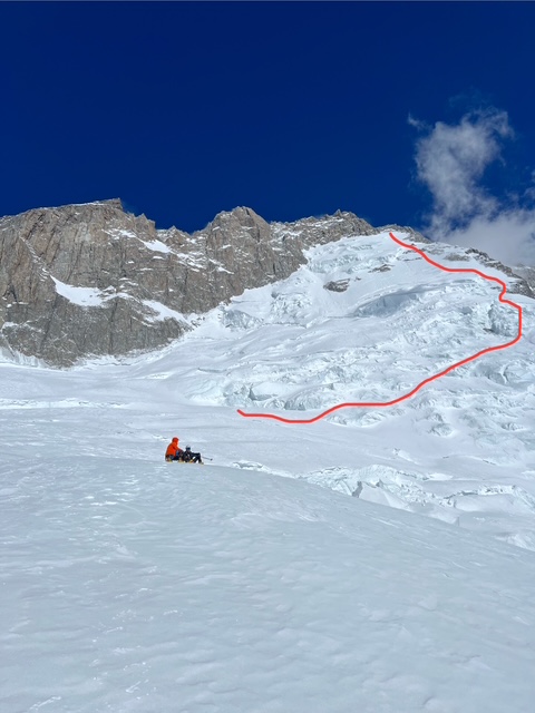 A climber sitting on the snow in front of a face, and a route across it marked in red