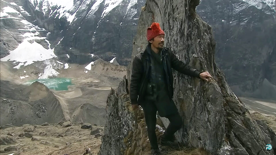 A man standing on a rocky mountainside, with snowy peaks in the distance.