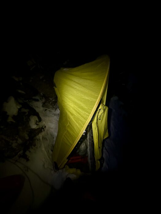 A hapf-pitched tent on a narrow snow ledge, lit by a headlamp in the night.