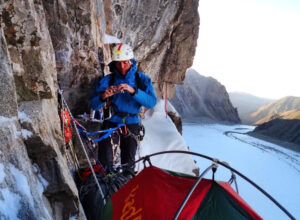 Pivtsov eats something while secured to an overhanging mixed face, his bivy tent at is feet