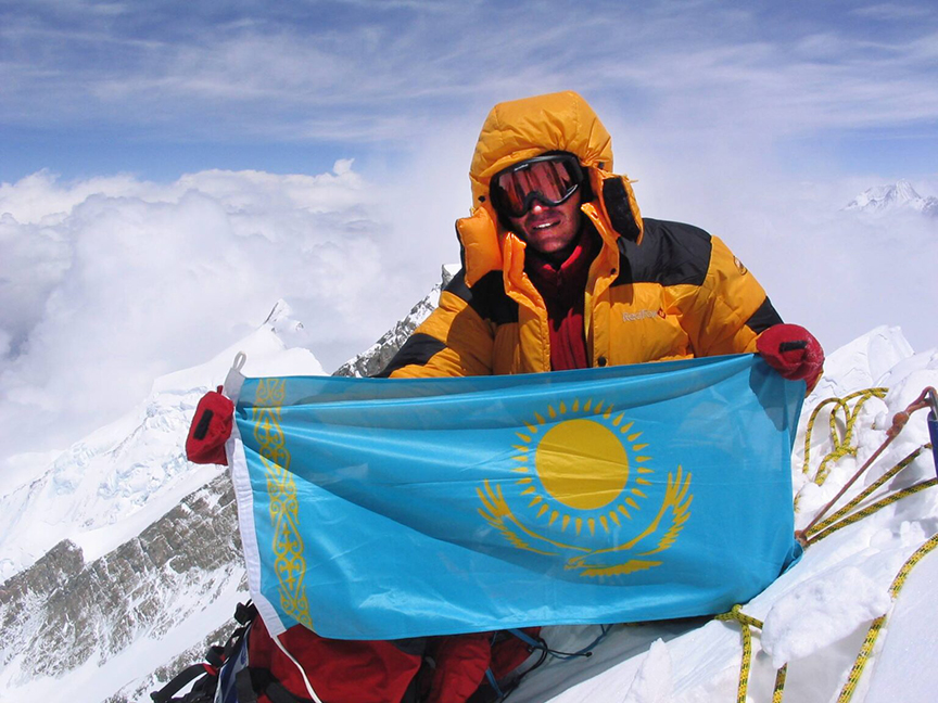 Pivtsov dressed in yellow down suit on a summit, holding Kazakhstan's flag.