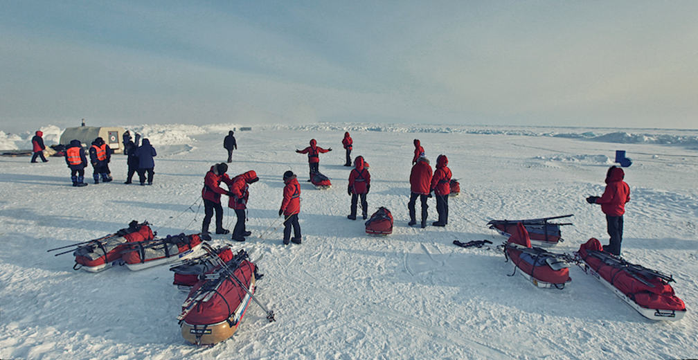 a bunch of people in red jackets standing on ice with quonset shelter in background