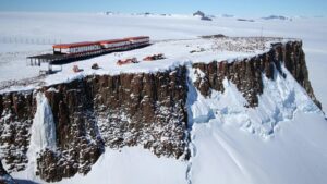 A series of three red painted buildings atop a rocky cliff in an Antarctic landscape.