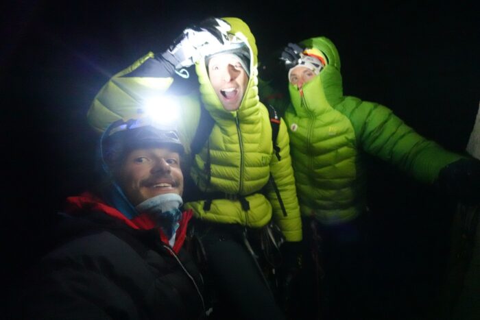 The climbers take a selfie on a Patagonia summit in pitch black night, with their headlamps and hoods on. 