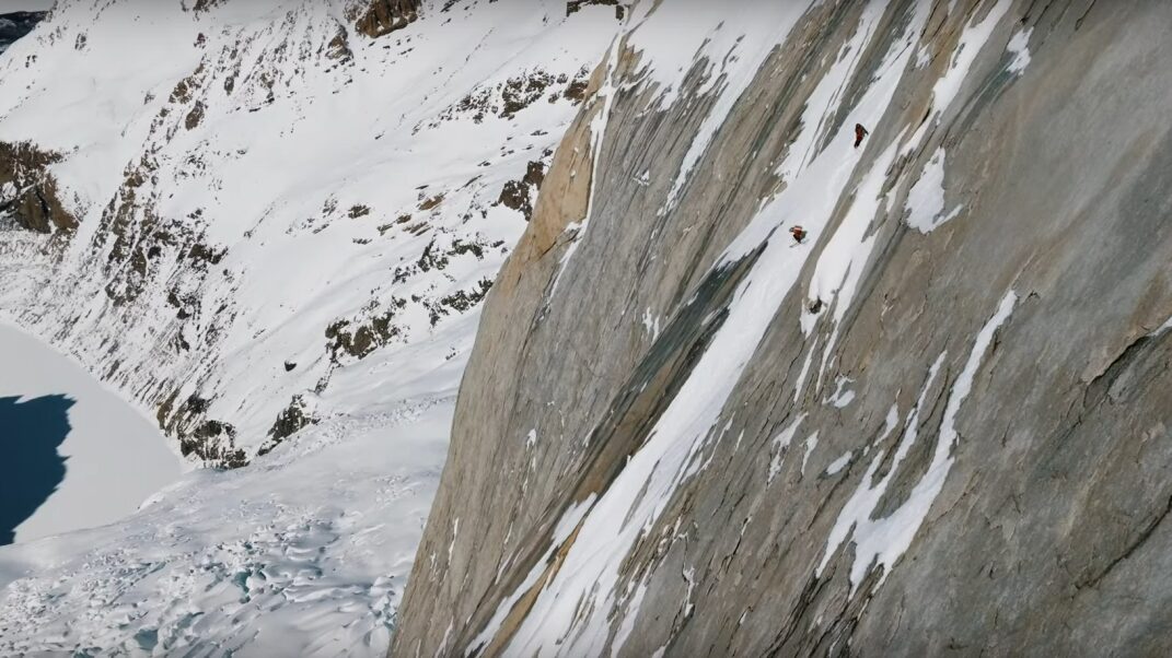 A sheer granite face with a thin band of snow, several figures can be seen sking down it.