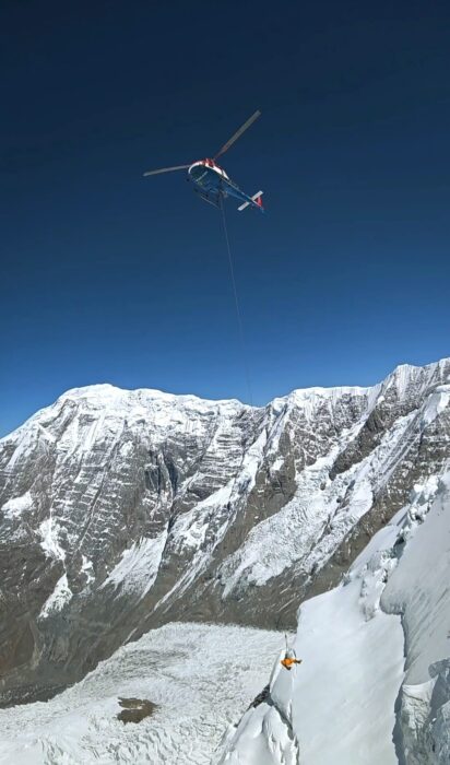 A helicopter above the peaks of Annapurna area, holding a climber at the end of a cable.