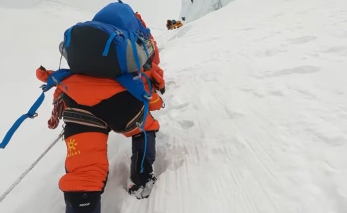 Climbers in high altitude gear climb on a steep snow section, clipped to a fixed rope.
