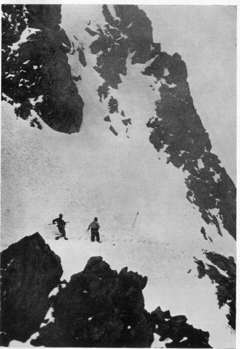 Two sherpas traverse a steep snow slope leaning on a pole, in a black and white image from 1963.