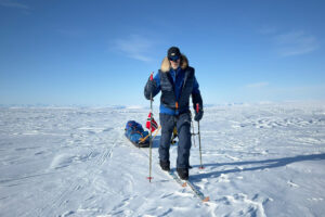 Borge Ousland dressed in winter gear skis across a vast, snowy Arctic landscape under a clear blue sky. He is pulling a sled loaded with supplies and a Norwegian flag attached. The terrain is icy and expansive, with distant mountains barely visible on the horizon.