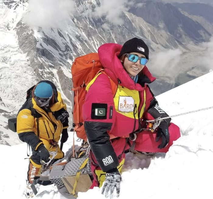 Naila Kiani smiles to the camera while climbing a steep snow slope clipped to a fixed rope.
