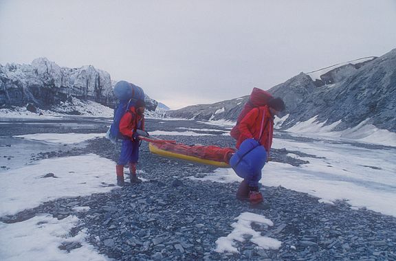 Two Arctic travelers in red jackets and blue pants pull a yellow sled loaded with gear across a rocky and snowy landscape. The terrain is rugged with patches of ice and gravel, flanked by snow-covered cliffs and mountains. The sky is overcast, and the scene conveys the difficulty of overland travel in the high Arctic.