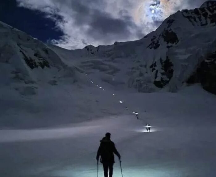 A line of people at dawn, lighted up by headlamps, heading up a snow-covered col in the Karakoram mountains. 