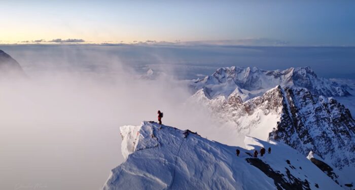A climber on the summit of Everest at sunrise, with a wind plume on the side of the moutnain.