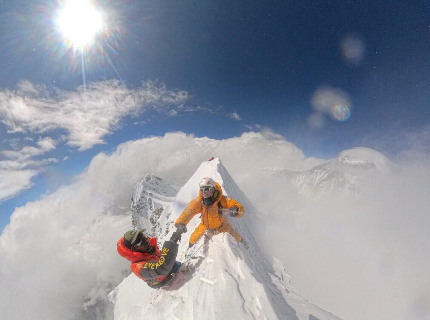 A climber sits on the edge of a sharp summit ridge and shakes hand with sherpa guide.