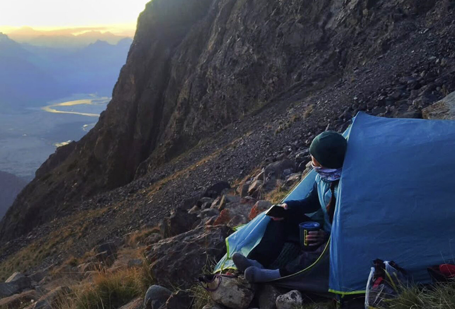 Cardell peers out from a tent pitched in a mountain slope, with no snow, at sunset.