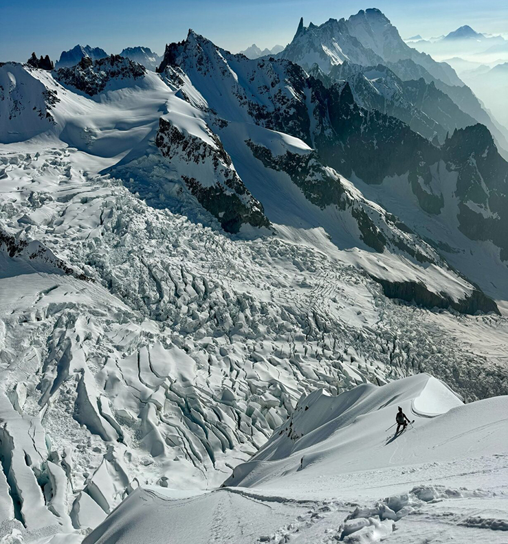 A skier on a snowy ridge on Mont Blanc, looks to the glacier below, very broken.