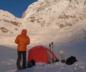A climber looking up a moutnain face from behind his tent on a snowy plane.
