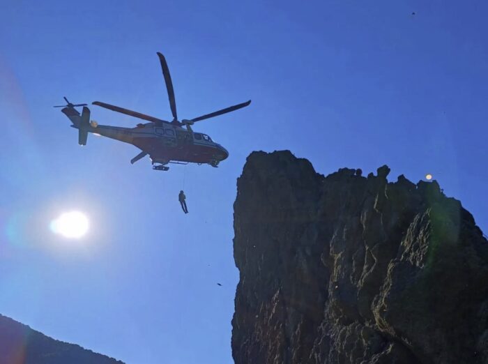A rescuer hangs from a helicopter on flight towards a rock wall.