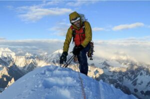 Chris Wright smiles on a spicky , snowy summit surrounded by mountain summits piercing the clouds.