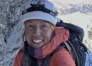Laura Dahlmeier smiles to the camera with climbing helmet and goetex jacket on, at a rick ledge on the north face of Eiger.