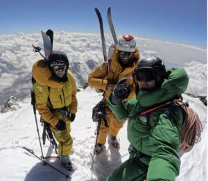 Three climbers on the snowy, flat summit of Nanga Parbat, to of them with skis on backpacks.