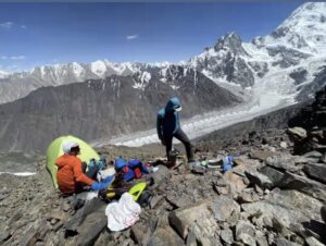 Two climbers in front of a small tent pitched on rocky morraine terrain.