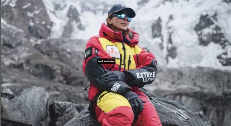Naila Kiani sits on a rick sourrounded by mountains covreed in clouds, and dressed in a dawnsuit.