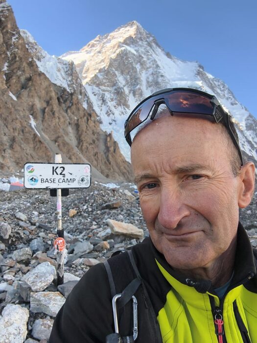 Close shot of Serge Hardy with a sign reading K2 BC and the mountain behind him.
