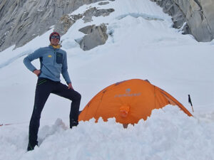 The climber standing by a semi buried tent in a snowy patch at the base of a granite wall.