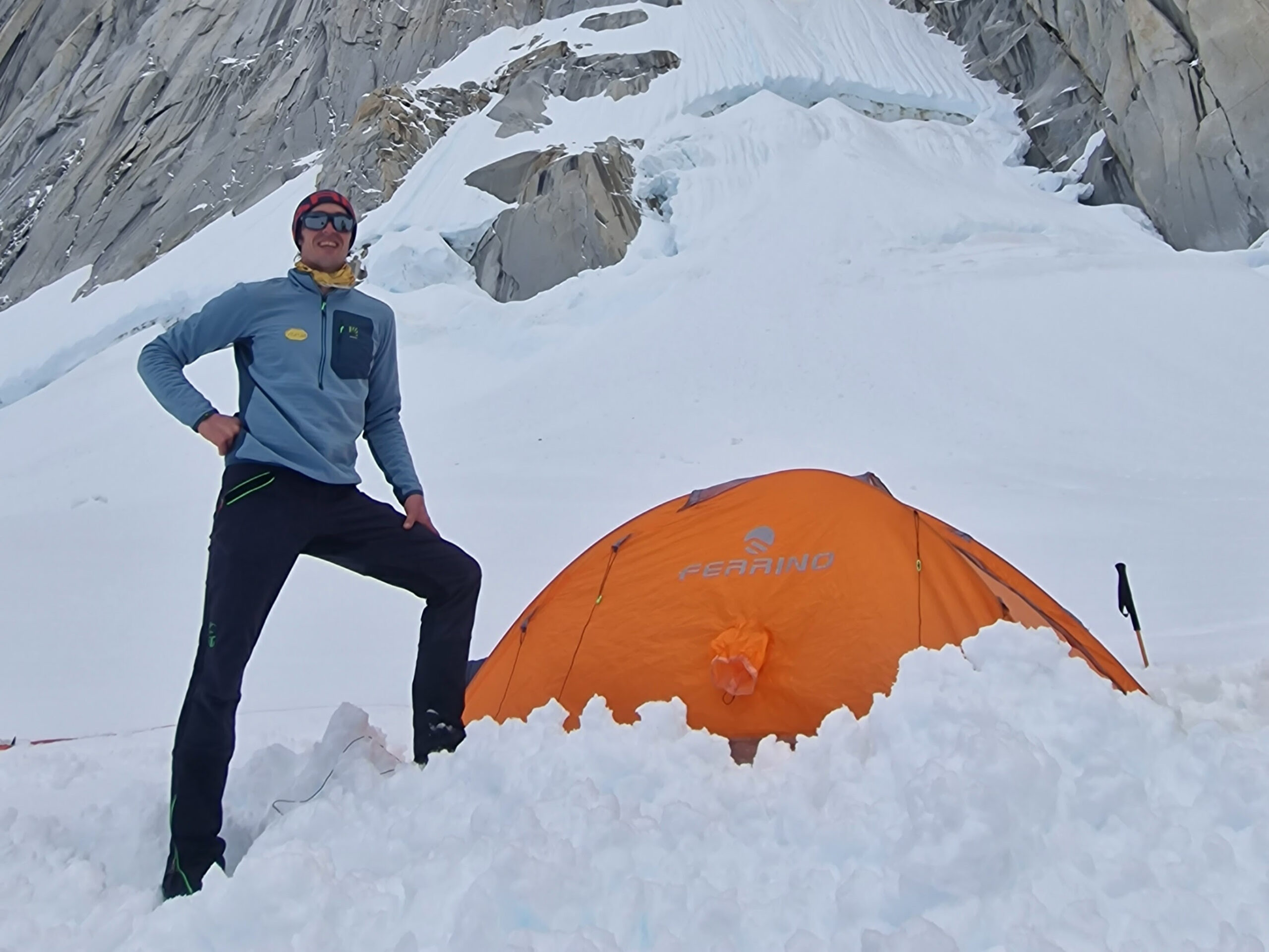 The climber standing by a semi buried tent in a snowy patch at the base of a granite wall.