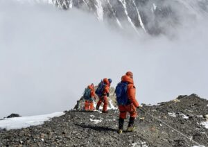 Three climbers descend on rocky ground clipped to a fixed rope, a cloud in front of them covers a mountain flank in background.
