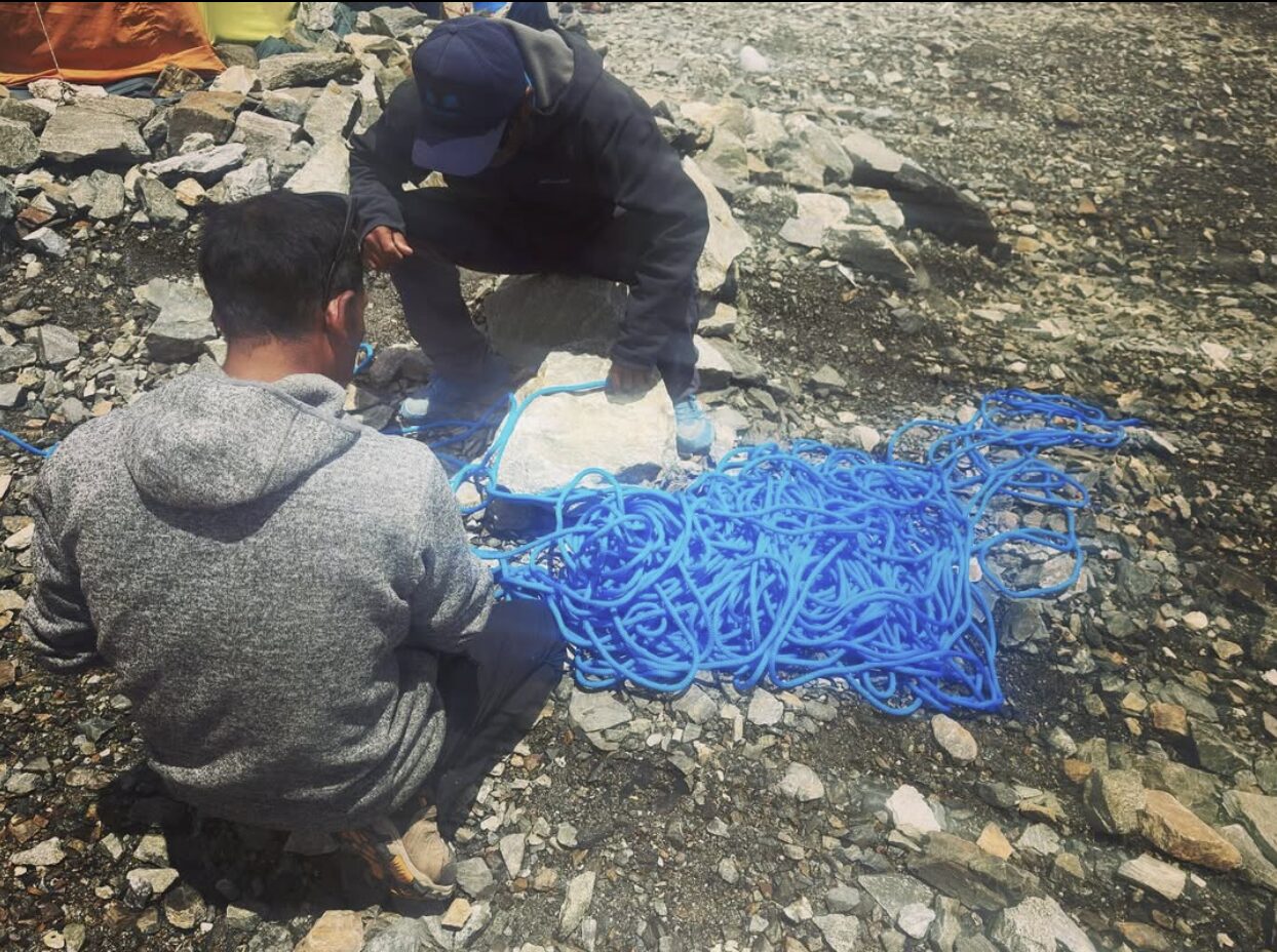 Two climbers squat at the rocky base camp and sort out oa maze of blue climbing rope.
