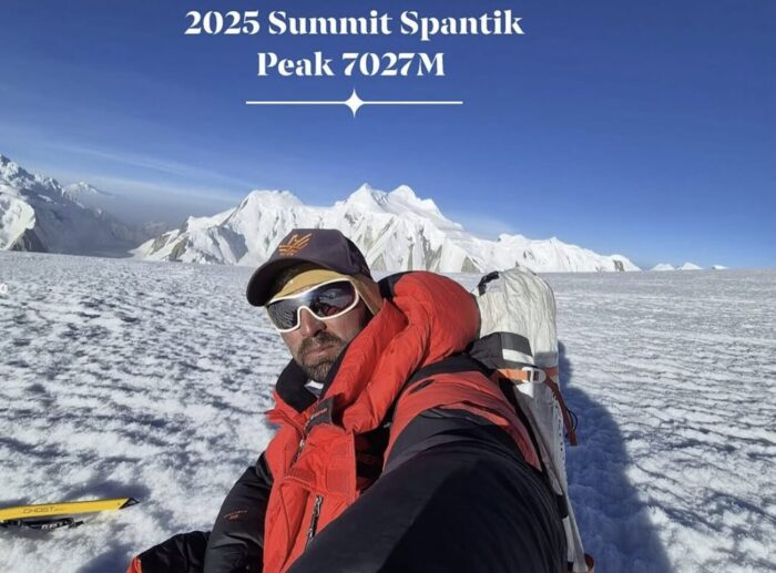 A climber takes a selfie while sitting on a snow plateau; summits in background.