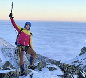 Bieleki rises his ice-axe while standing on a mixed ridge with a sea of clouds in background below.