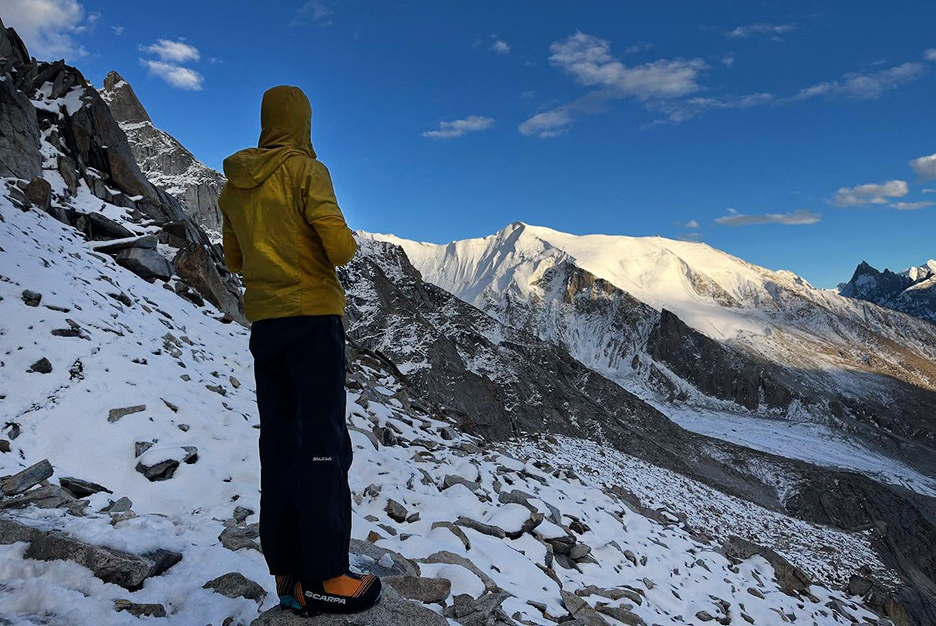 A climber in shade looks toward a mountain with the summit lit by the morning sunrays.