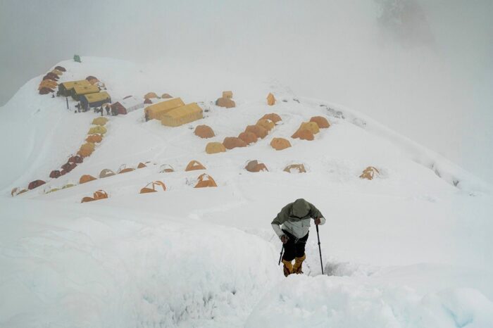 Carlos soria in a cloudy day, heads up a snowy trail with the tents of Camp 1 behind him. 