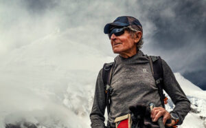 Close shot of Carlos Soria in t-shirt, cap and sunglasses. Manaslu's summits appears in background, wrapped in clouds.