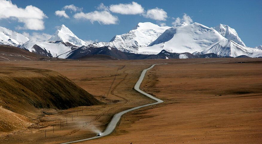 A road on a brown plane leads to stunningly white, snow-covered mountains in background.