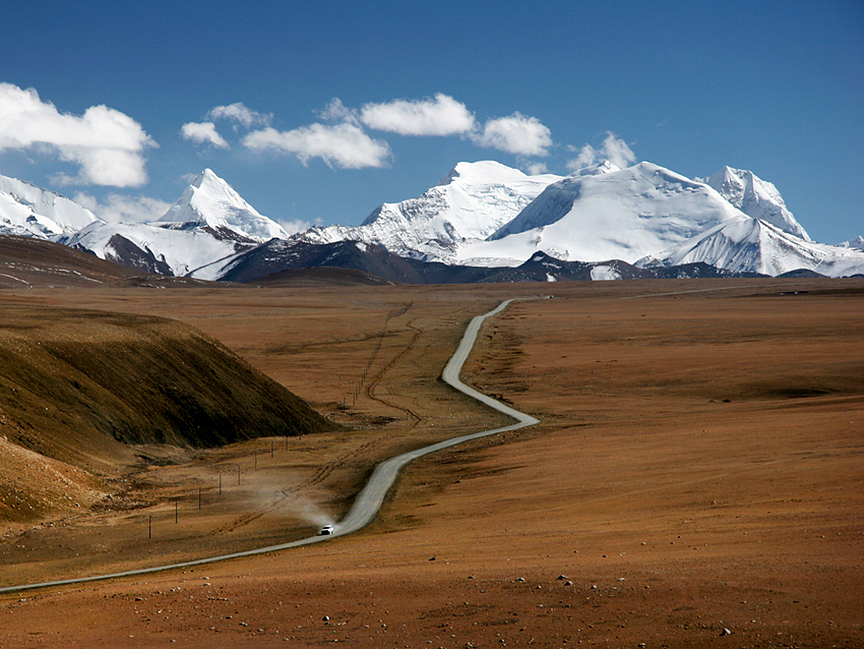 A road on a brown plane leads to stunningly white, snow-covered mountains in background.