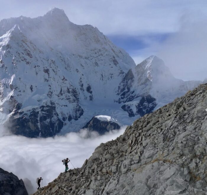 Two climbers on the edge of a rocky ridge, with the north face of Jannu in background.