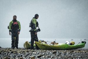 Two kayakers stand next to their loaded boats in the pouring rain