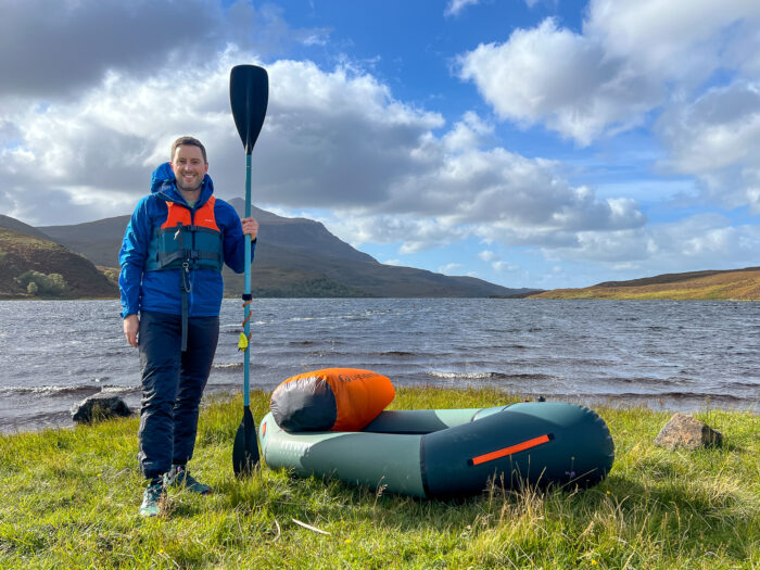 A man stands next to a packraft holding a paddle, with a loch in the background