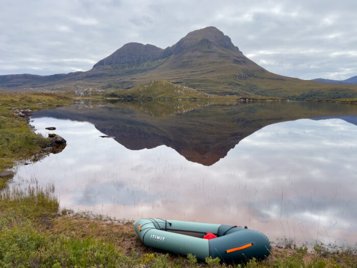 A packraft sits in front of a loch reflecting a mountain in the background