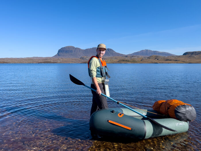 A man stands in shallow water of a loch with a packraft next to him
